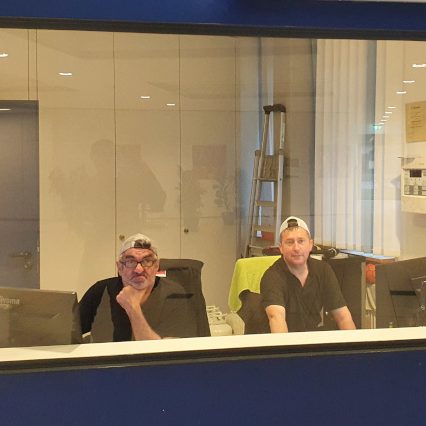 Hausmeister Zwei Männer sitzen an Schreibtischen in einem Büro, Blick durch eine Glaswand.