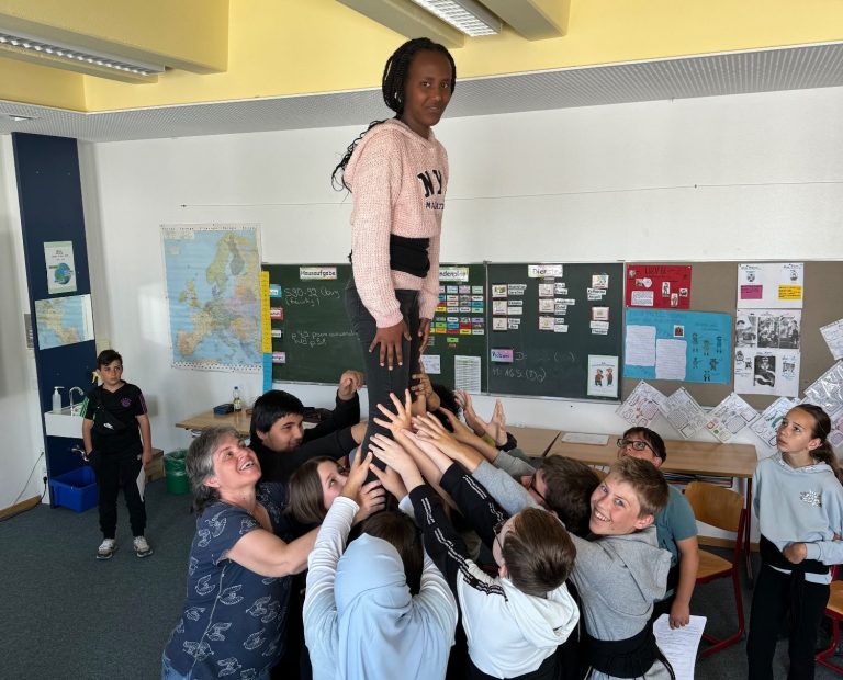 Erasmus+ Gruppenfoto Ein Kind steht auf einem Haufen anderer Kinder, die sie von unten stützen.
